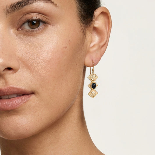 A close-up of a model's ear wearing a gold drop earring with three stacked geometric charms: a gold oval, a gold diamond with a clear stone, and a central round dark blue stone.