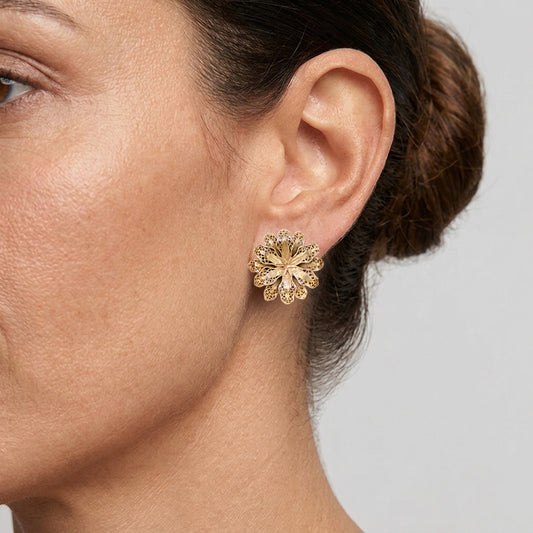 A close-up of a woman's ear wearing a textured gold flower-shaped stud earring against a light gray background.