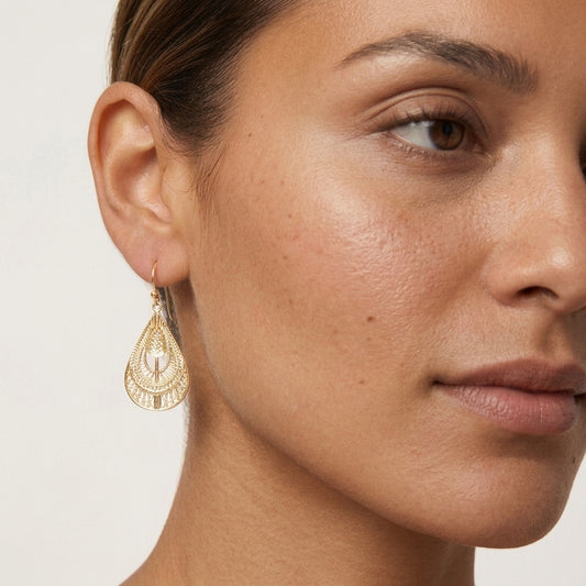 A close-up of a woman's ear and side profile wearing a delicate gold teardrop dangle earring with an intricate filigree pattern.
