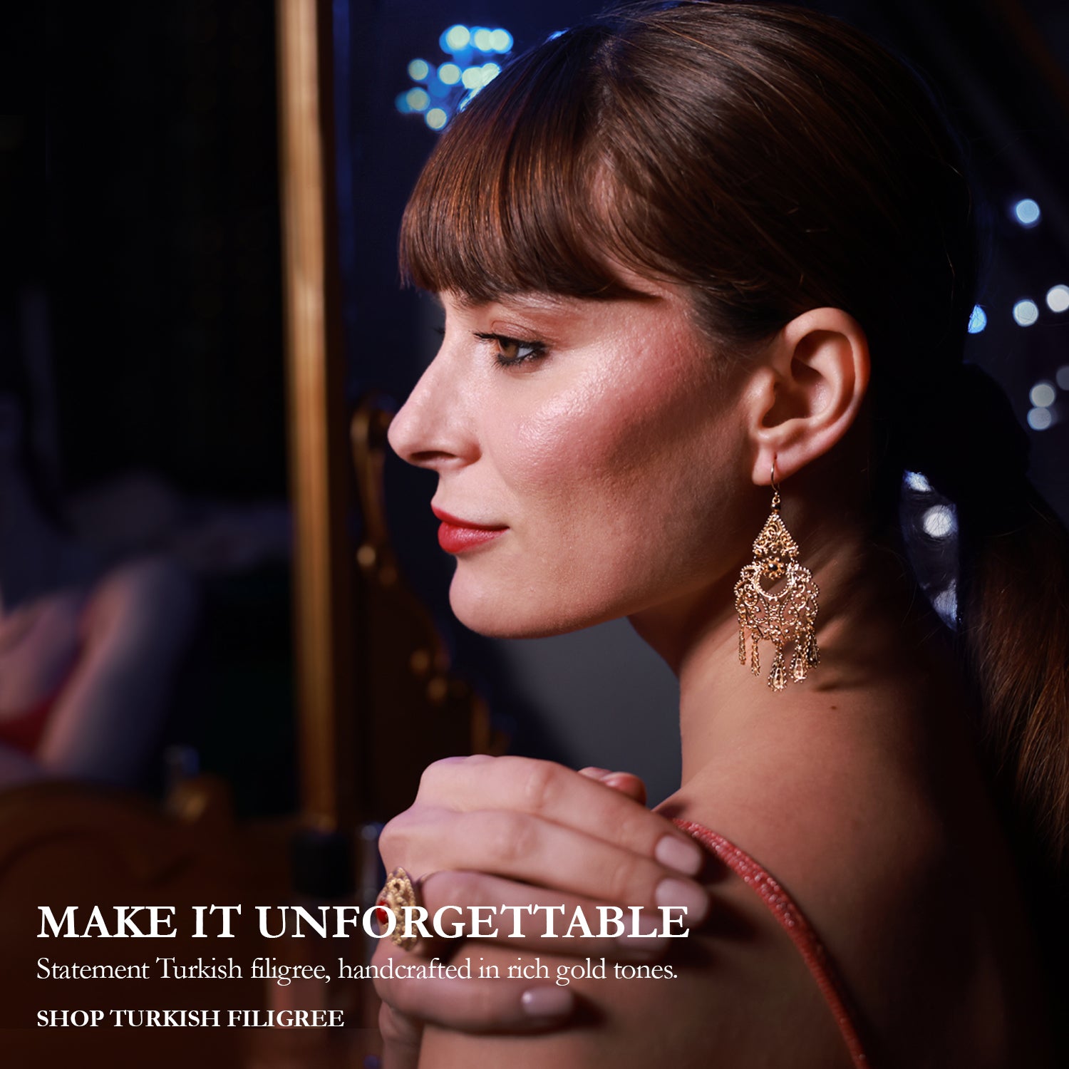 A woman in profile wearing ornate gold statement earrings and a ring, with her hands clasped in front of a mirror showing the back of the earrings. The image has a dark, elegant background with bokeh lights and text on the left reading 'MAKE IT UNFORGETTABLE' and 'Statement Turkish filigree, handcrafted in rich gold tones.