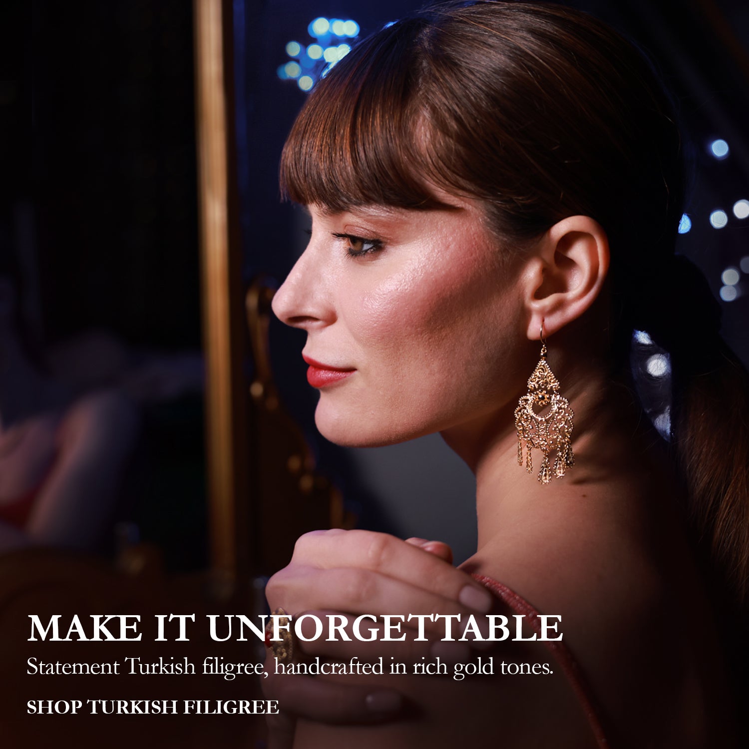 A woman in profile wearing ornate gold statement earrings and a ring, with her hands clasped in front of a mirror showing the back of the earrings. The image has a dark, elegant background with bokeh lights and text on the left reading 'MAKE IT UNFORGETTABLE' and 'Statement Turkish filigree, handcrafted in rich gold tones.
