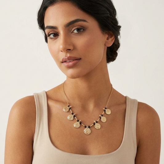 A woman wearing a gold coin pendant necklace with small black bead accents, styled with a beige tank top against a light off-white background.