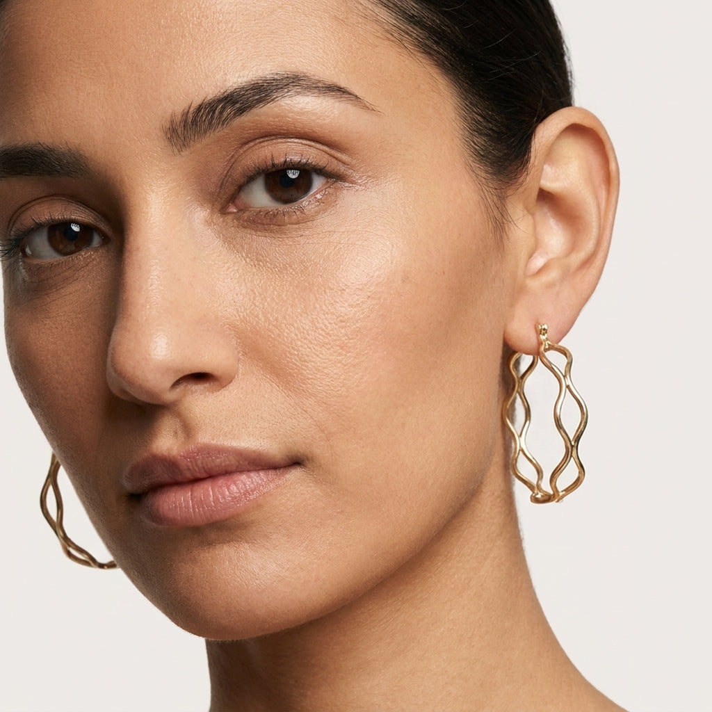 A close-up of a model wearing a pair of gold-toned, twisted open hoop earrings against a light gray background.
