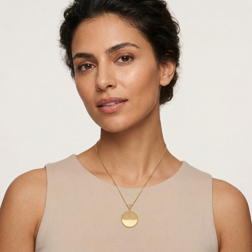 A woman wearing a delicate gold chain necklace with a textured circular pendant, styled with a beige sleeveless top against a clean off-white background.
