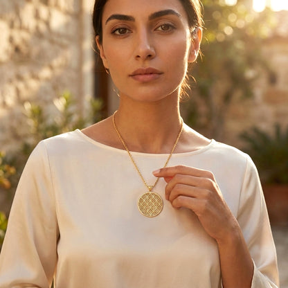 A woman wearing a delicate gold chain necklace with a round, textured gold pendant, posing outdoors against a warm, sunlit stone wall background.