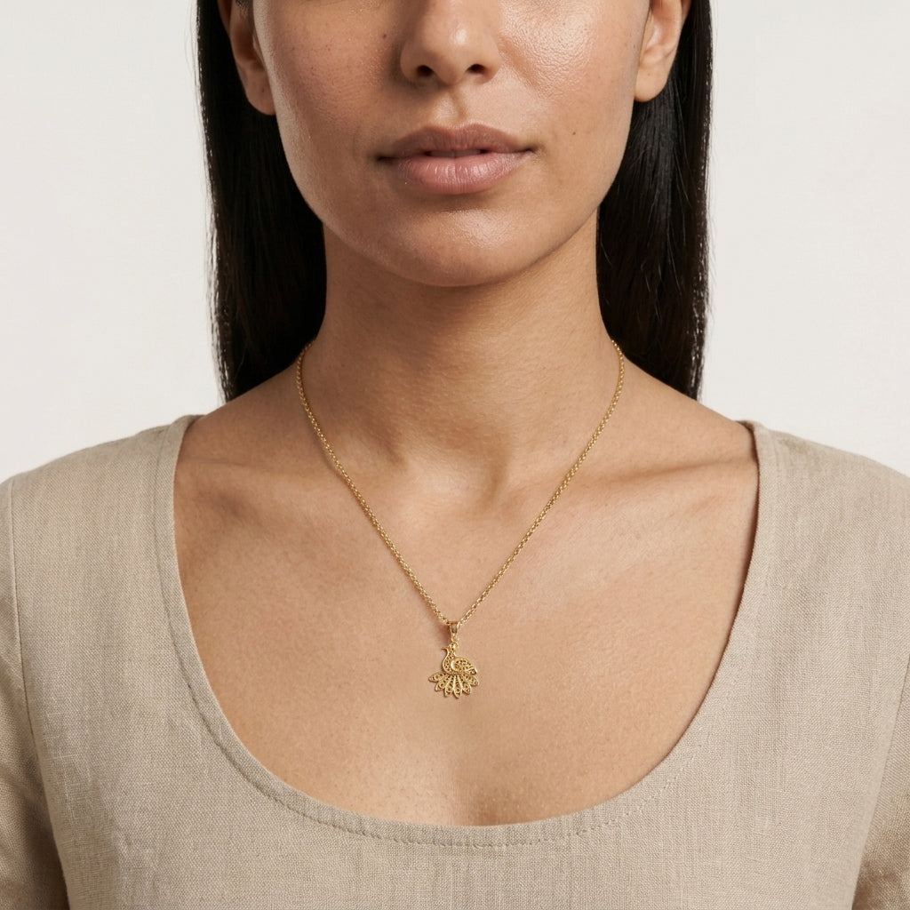 A close-up of a woman wearing a delicate gold chain necklace with a small, textured gold pendant, paired with a beige linen top against a light off-white background.