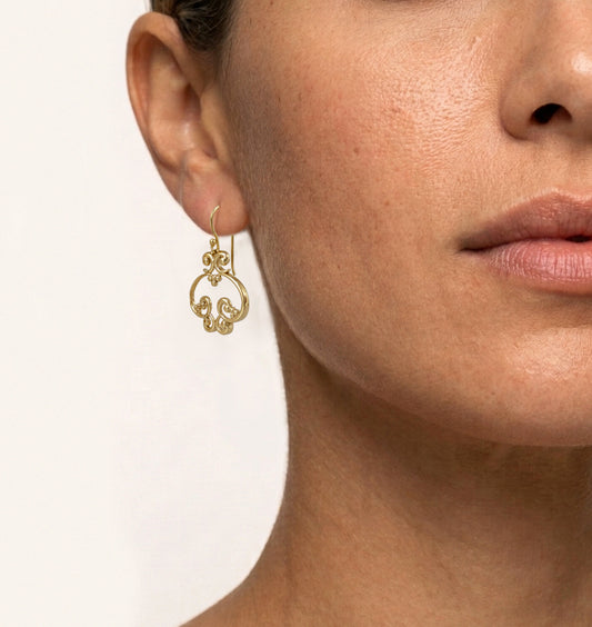 A close-up of a model's ear and neck wearing a delicate gold hoop earring with a small ornate dangling charm, against a clean off-white background.
