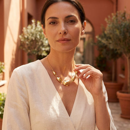 A woman wearing a chunky gold statement necklace with organic-shaped links, styled with a white linen wrap top in a warm, sunlit outdoor setting.