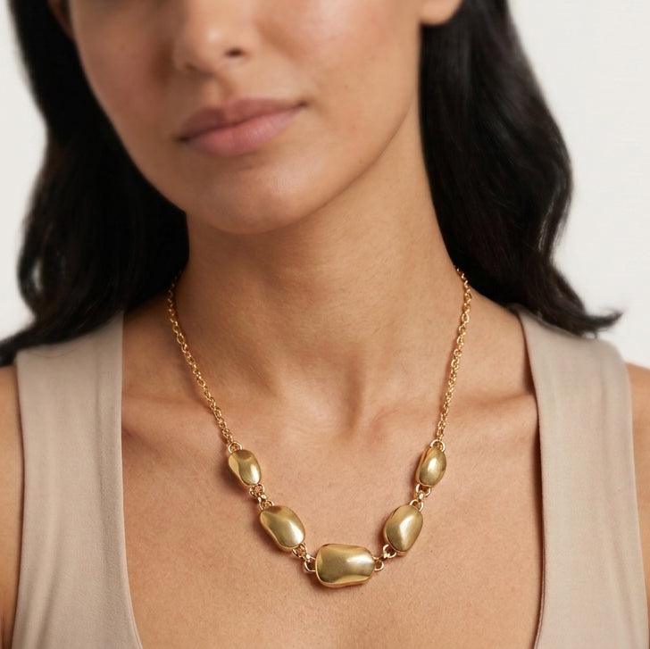 A close-up of a woman wearing a gold necklace with irregular, polished stone-like beads on a delicate chain, paired with a beige sleeveless top.