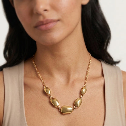 A close-up of a woman wearing a gold necklace with irregular, polished stone-like beads on a delicate chain, paired with a beige sleeveless top.