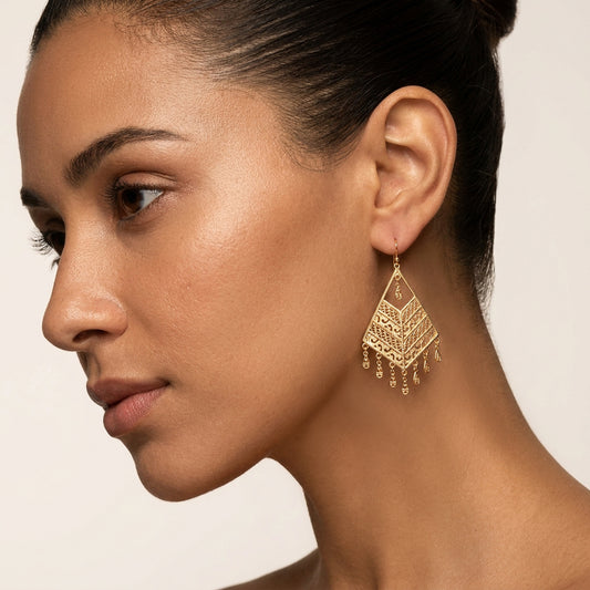 A close-up side profile of a woman wearing a large gold-toned geometric dangle earring with dangling chain details against a plain light beige background.
