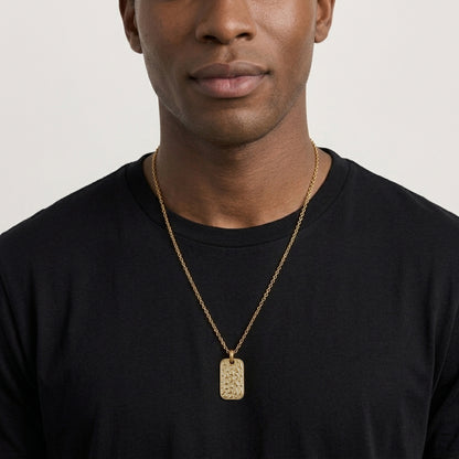 A close-up of a man wearing a textured gold rectangular pendant necklace on a gold chain over a black t-shirt, against a light gray background.