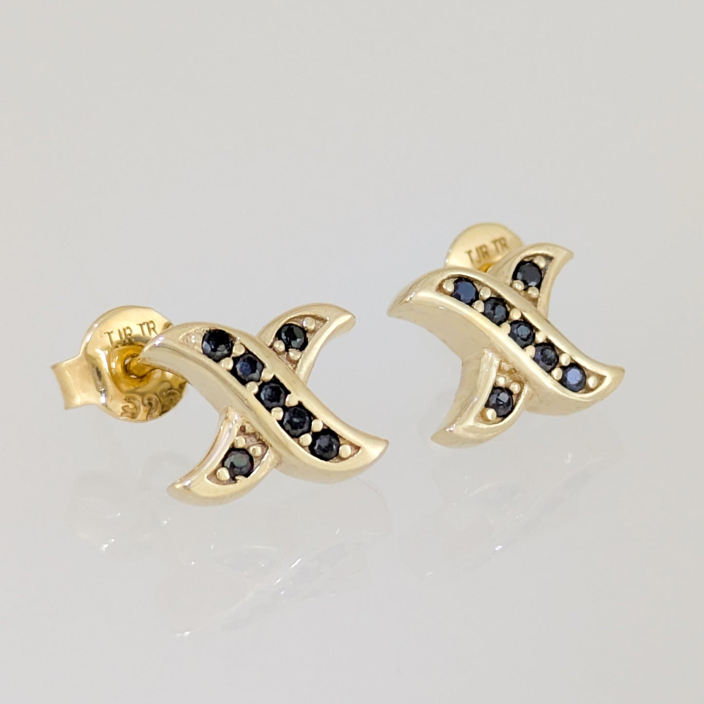 A pair of gold-toned stud earrings with a curved, ribbon-like design set with small dark  gemstones, shown on a light gray reflective surface.