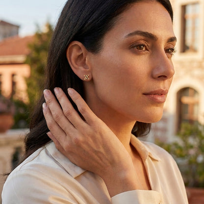 A woman wearing a cream-colored blouse and small, X-shaped gold earrings stands against a warm, softly blurred outdoor backdrop.