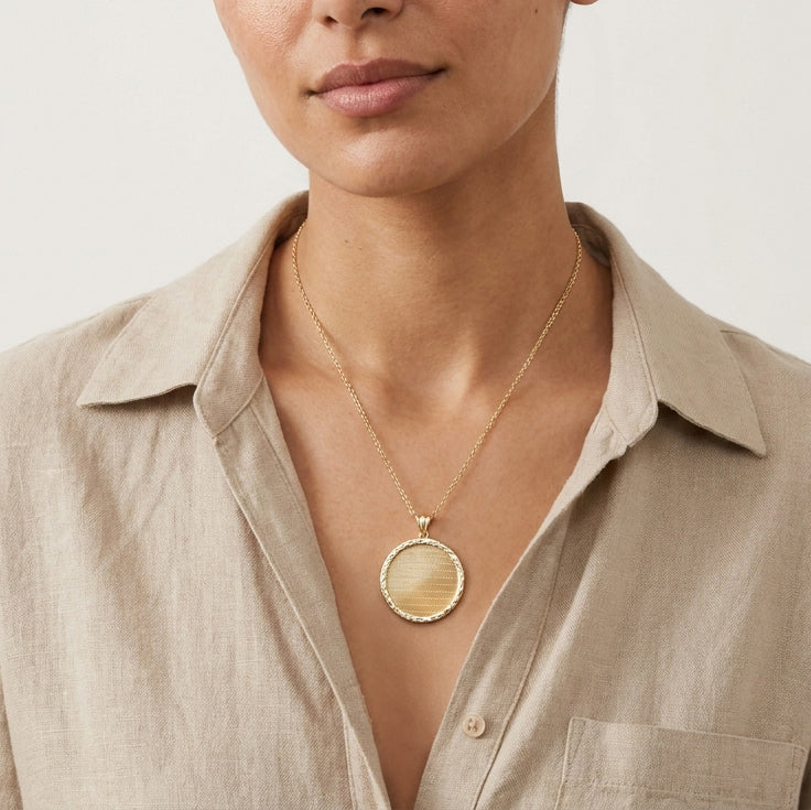 A close-up of a model wearing a gold pendant necklace with a circular, textured gold medallion, styled with a beige linen button-up shirt.