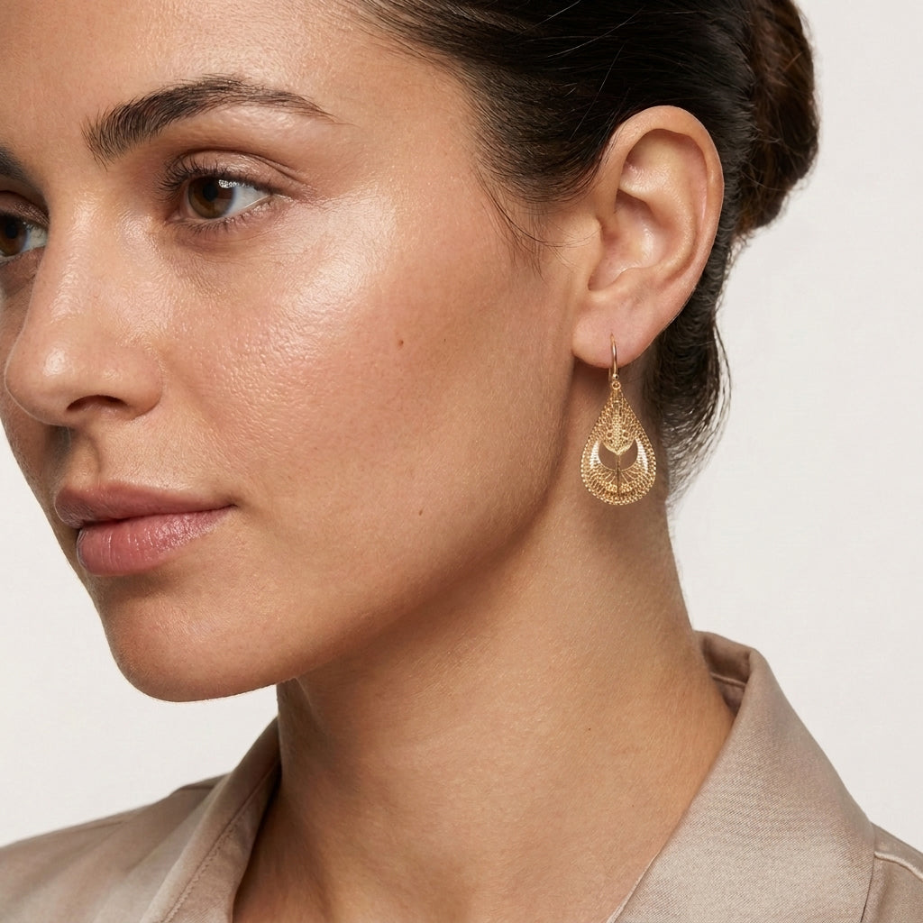 A close-up of a woman wearing a delicate gold teardrop dangle earring with an intricate openwork pattern, set against a clean, light off-white background.