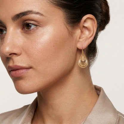A close-up of a woman wearing a delicate gold teardrop dangle earring with an intricate openwork pattern, set against a clean, light off-white background.