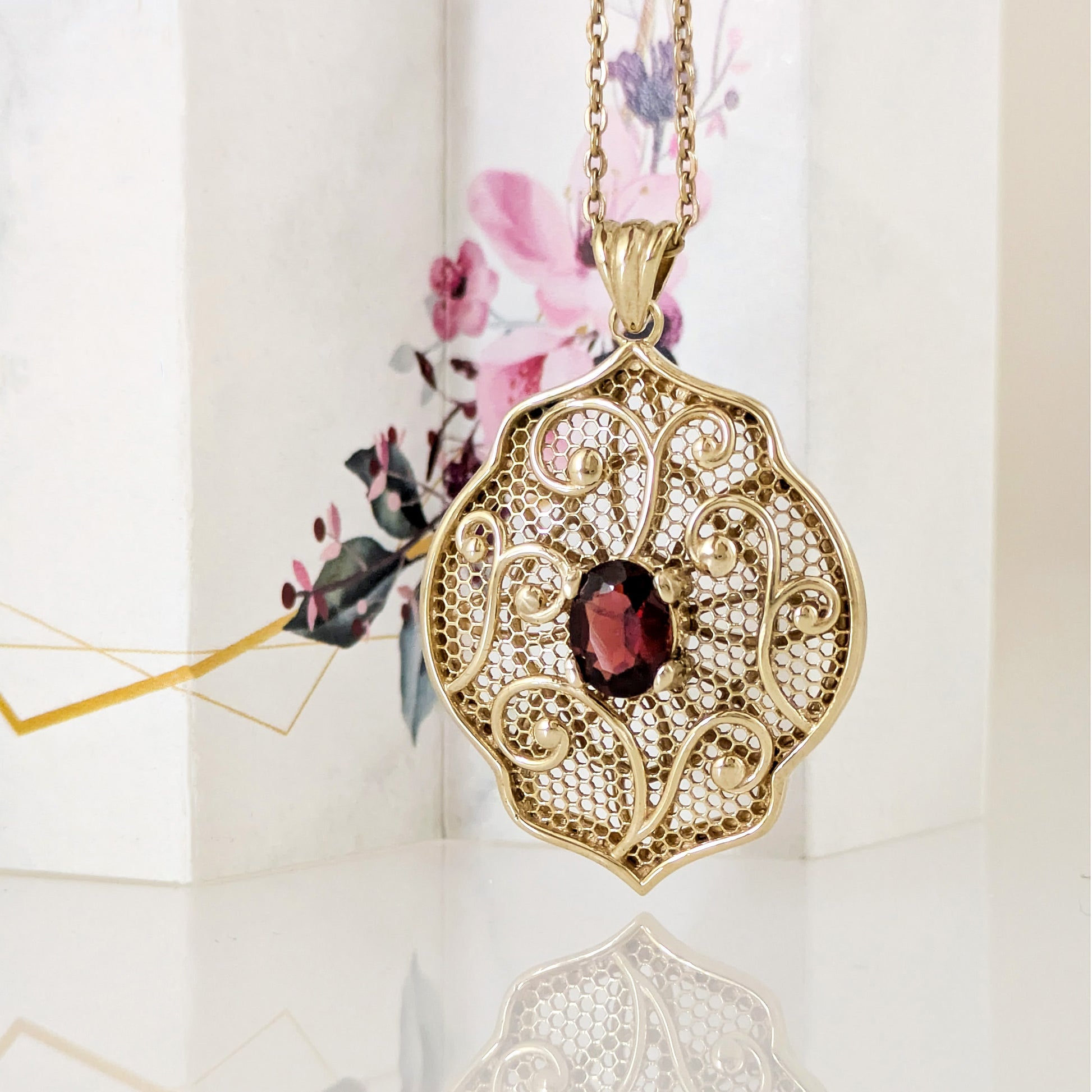 A gold filigree pendant necklace with an ornate openwork design and a central dark red oval gemstone, displayed against a soft white background with a floral illustration.
