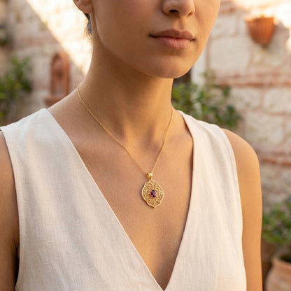 A close-up of a woman wearing a delicate gold pendant necklace with an ornate filigree design and a small red gemstone in the center, paired with a white sleeveless top.