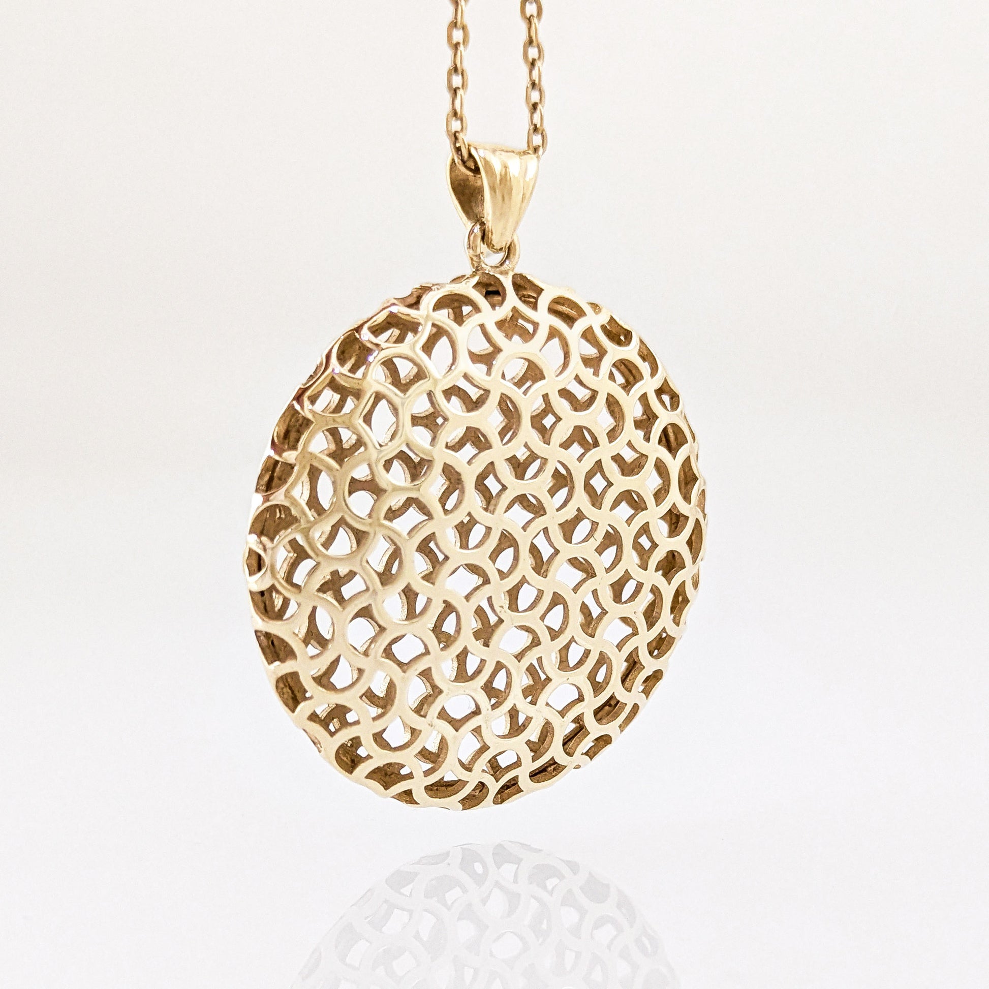 A gold pendant necklace featuring an intricate, openwork geometric mesh pattern on an oval pendant, suspended from a matching chain against a clean white background.