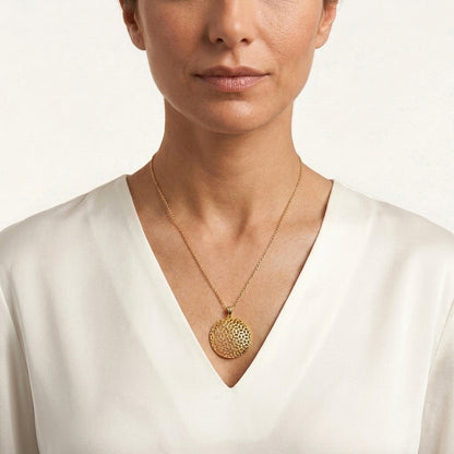 A close-up of a woman wearing a delicate gold chain necklace with a textured, circular gold pendant, styled with a white V-neck top against a clean white background.