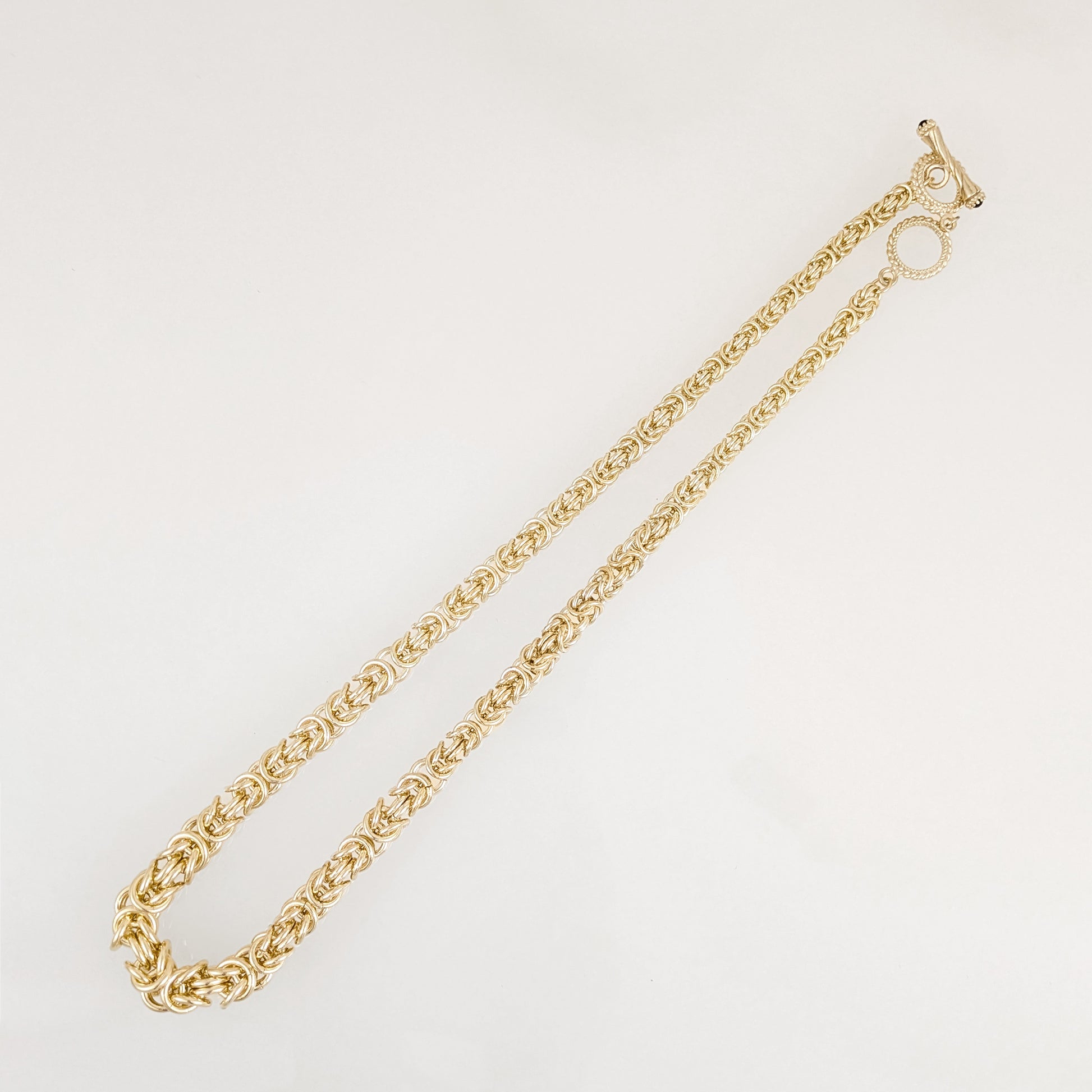 A gold graduated byzantine chain necklace with a distinctive textured, rope-like link pattern and a T-bar clasp with black onyx, displayed on a clean white background.