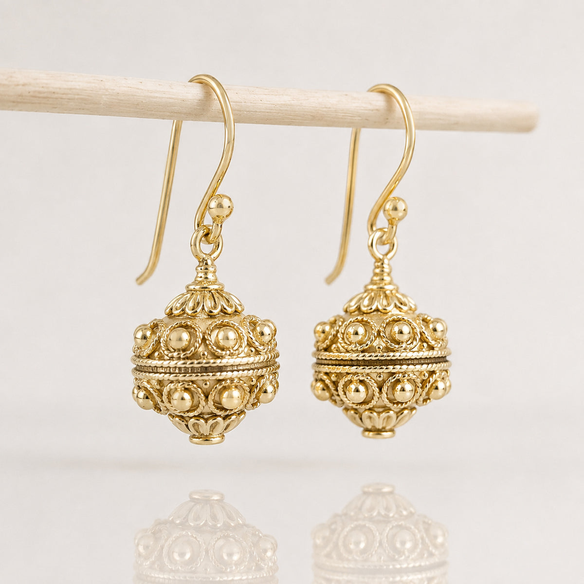 A pair of ornate gold-toned dangle earrings with intricate filigree detailing and small bead accents, hanging from a light wooden rod against a clean white background.