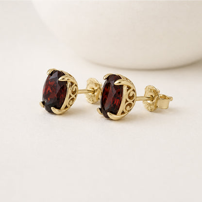 A pair of gold stud earrings with oval dark red gemstones, displayed on a clean white surface with a soft-focus white ceramic object in the background.