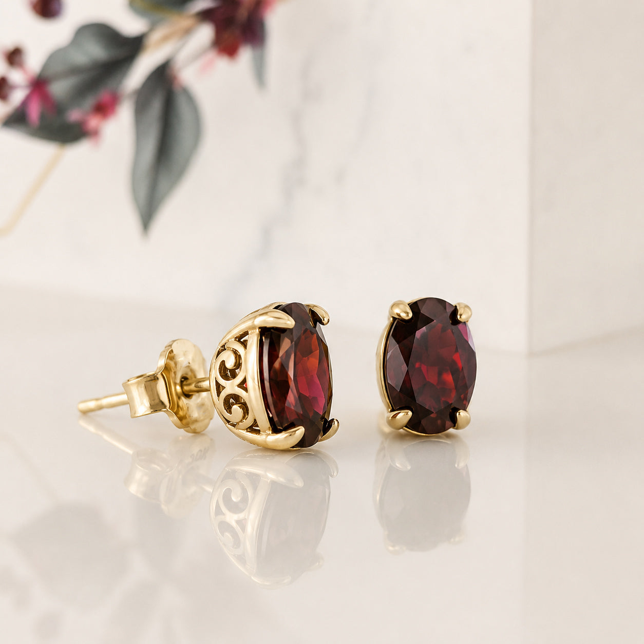 A pair of gold stud earrings with oval dark red gemstones, one shown from the front and the other from the side/back. They are displayed on a reflective white surface with a soft-focus background of marble and leaves.