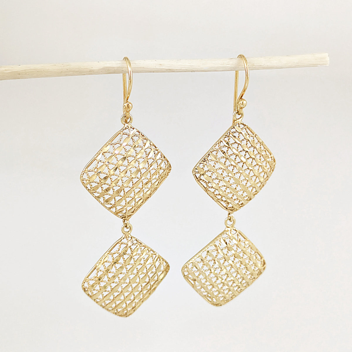 A pair of gold drop earrings with textured, openwork diamond-shaped pendants hanging from gold fishhook wires, displayed against a light gray background on a thin wooden stick.