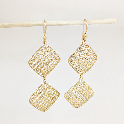A pair of gold drop earrings with textured, openwork diamond-shaped pendants hanging from gold fishhook wires, displayed against a light gray background on a thin wooden stick.