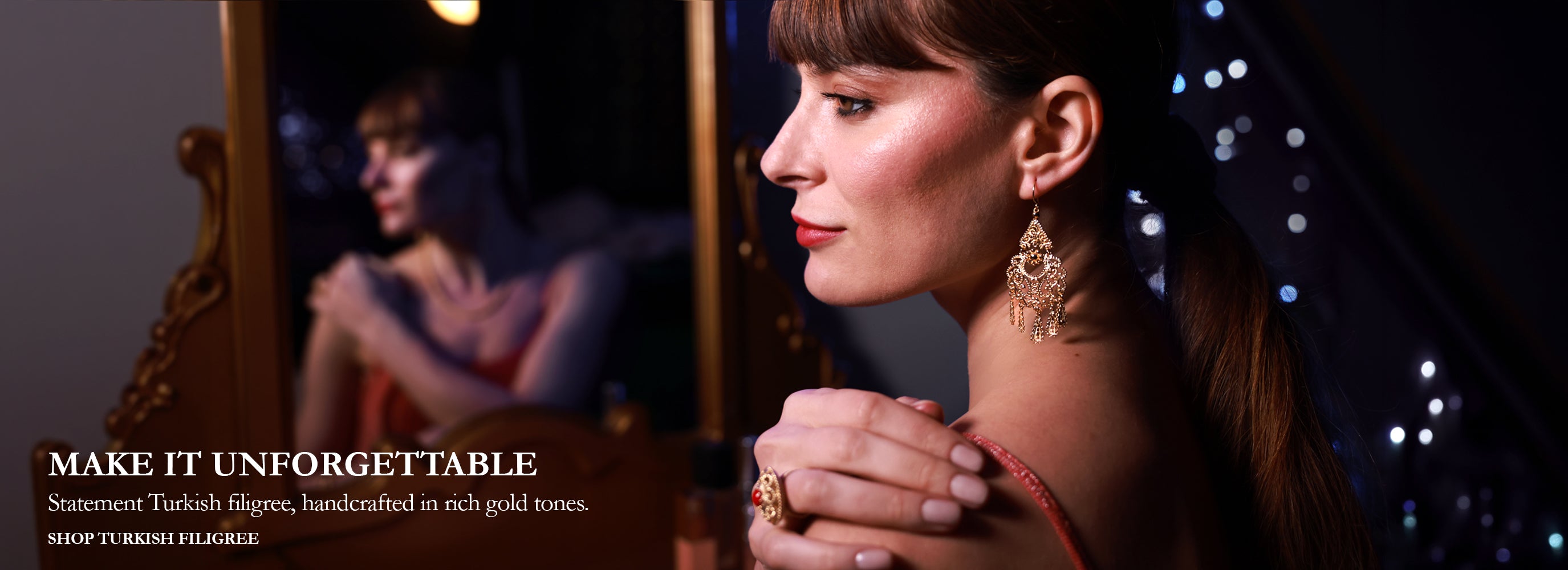 A woman in profile wearing ornate gold statement earrings and a ring, with her hands clasped in front of a mirror showing the back of the earrings. The image has a dark, elegant background with bokeh lights and text on the left reading 'MAKE IT UNFORGETTABLE' and 'Statement Turkish filigree, handcrafted in rich gold tones.