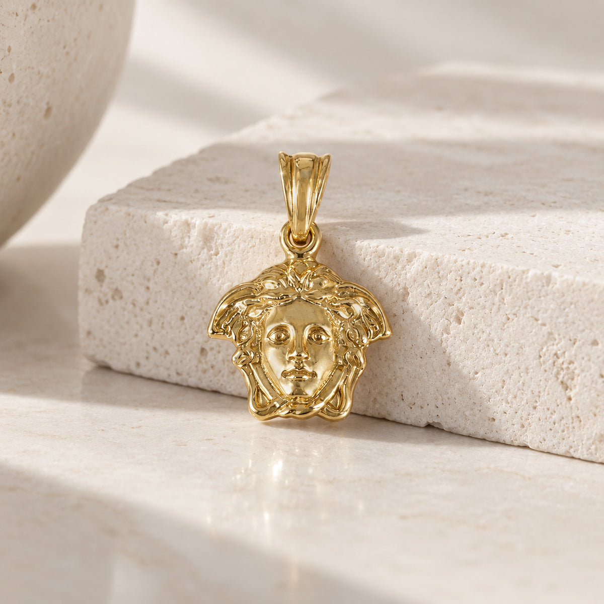 A gold pendant featuring a Medusa head design, displayed on a textured white stone block with soft shadows.