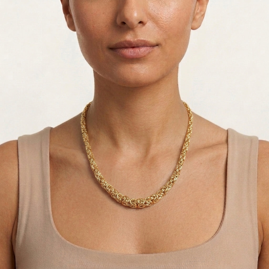 A close-up of a woman wearing a gold graduated byzantine chain necklace with an intricate woven or braided design, paired with a beige tank top against a light neutral background.